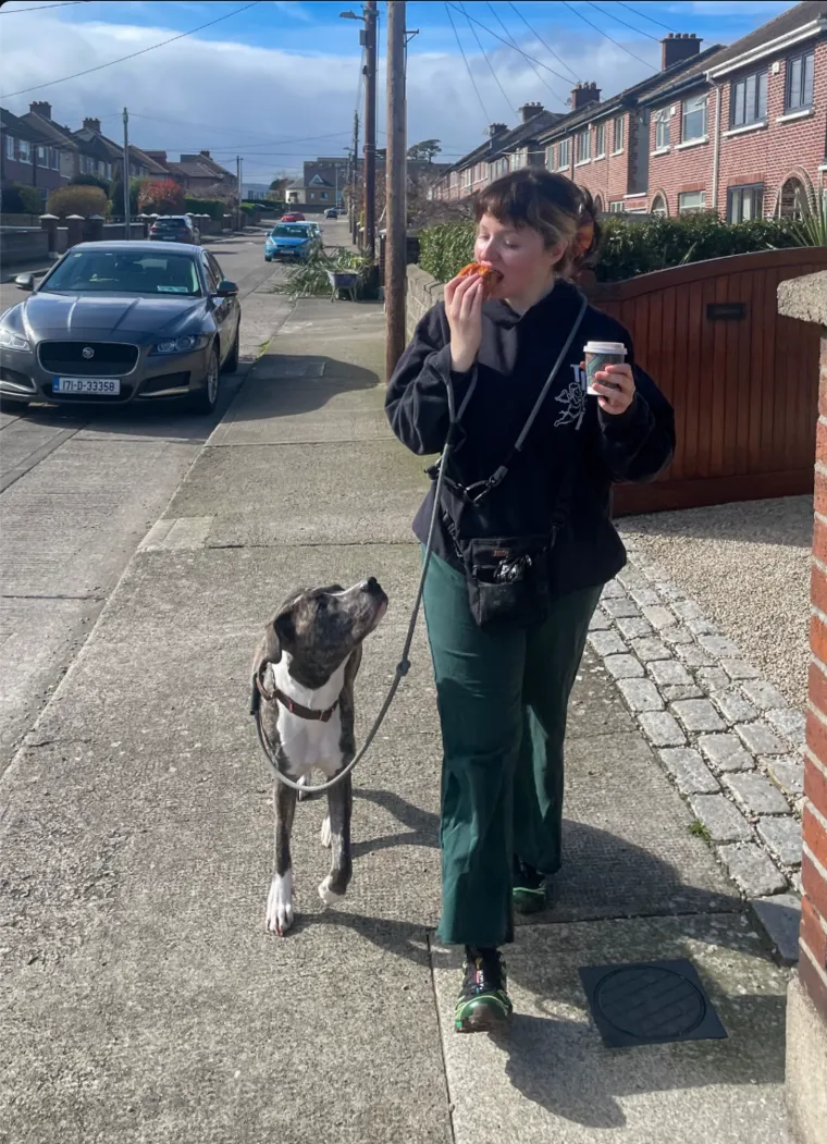 Sian eating a croissant while Goose walks beside her on a loose leash, looking up