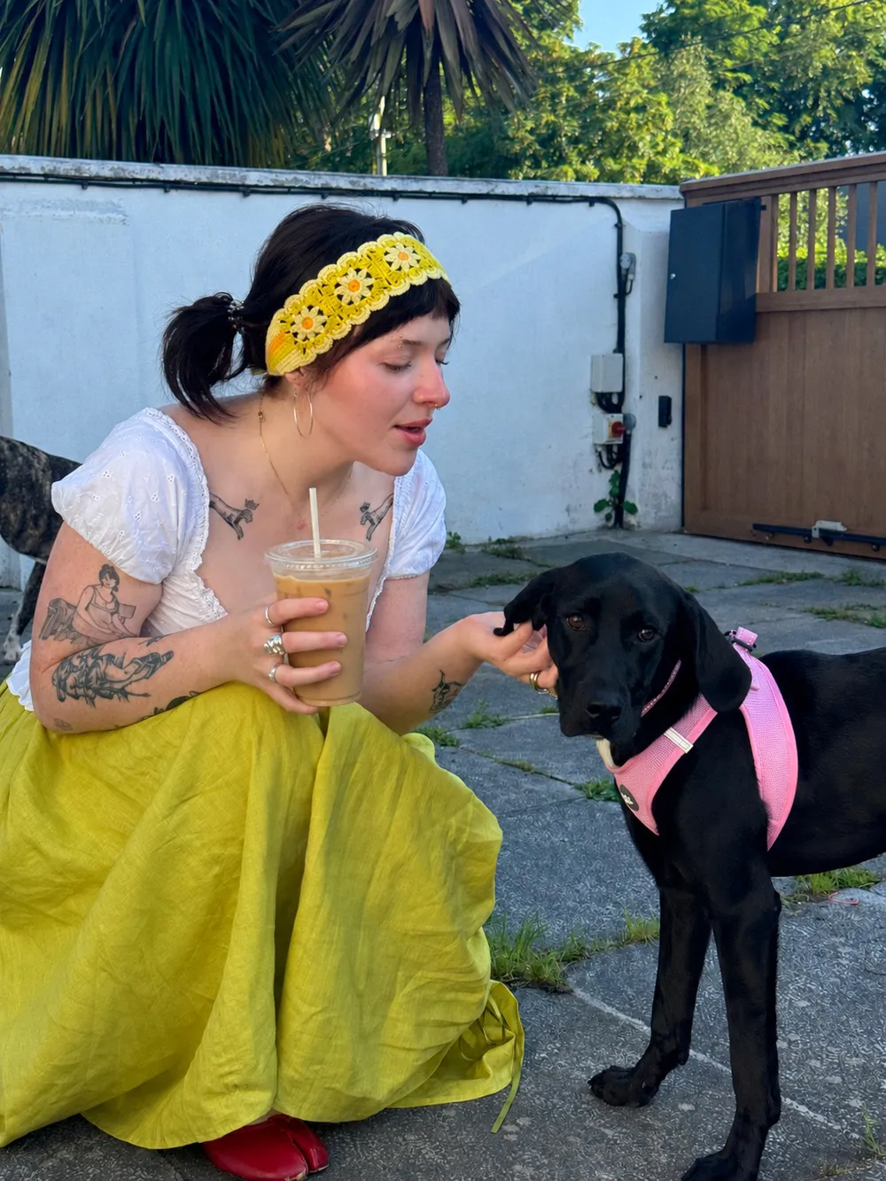 Sian crouching with an iced coffee, scratching Púca, a black puppy in a pink harness